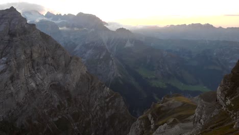 Aerial-view-of-the-cliffs-and-mountains-of-Linthal-in-Glarus,-Switzerland-during-sunset-with-pan-up-from-valley-to-mountain-peaks-with-glaciers