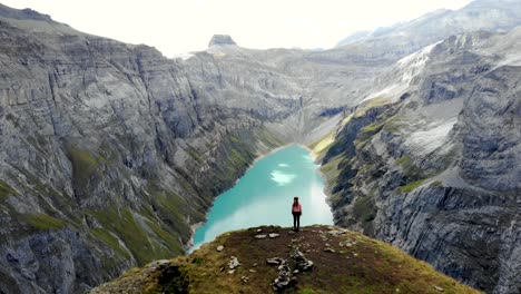 A-standing-hiker-enjoying-the-view-over-lake-Limmernsee-in-Glarus,-Switzerland,-the-turquoise-colored-water-of-which-is-surrounded-by-tall-Swiss-Alps-peaks-and-cliffs