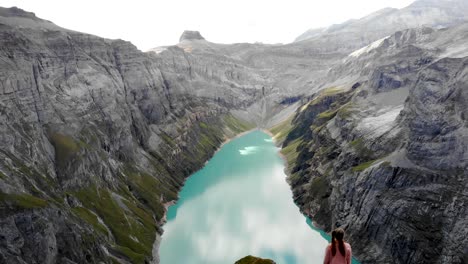 A-reverse-flyover-above-a-hiker-enjoying-the-view-over-lake-Limmernsee-in-Glarus,-Switzerland,-the-turquoise-colored-water-of-which-is-surrounded-by-tall-Swiss-Alps-peaks-and-cliffs