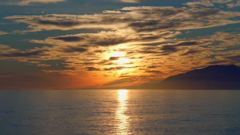 Dramatic-sunset-with-calm-ocean-waves,-clouds,-and-mountains-on-the-background