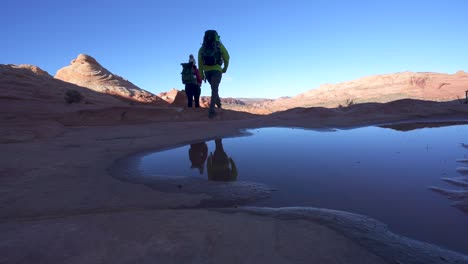 Two-hikers-wearing-backpacks-walk-past-pool-of-water-in-rocky-Desert