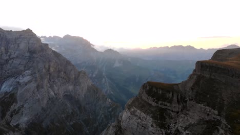 Aerial-view-of-the-cliffs-and-mountains-of-Linthal-in-Glarus,-Switzerland-during-sunset-with-pan-down-from-mountain-peaks-with-glaciers-to-the-canyon-like-valley