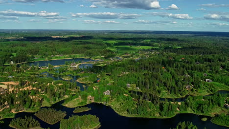 Drohne-Fliegt-über-Die-Malerische-Landschaft-Von-Amatciems,-Ein-Lettisches-ökodorf,-Das-Durch-Einen-See-Und-üppige-Grüne-Bäume-Geschnitzt-Ist