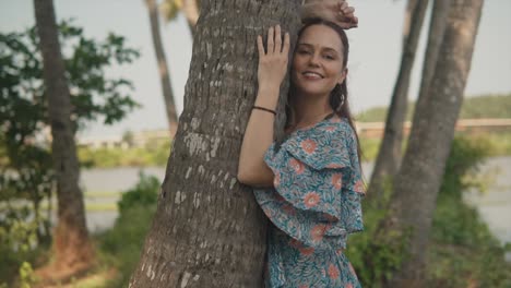 Beautiful-young-woman-in-a-dress-leaning-on-a-tree-smiling-with-one-hand-on-her-head