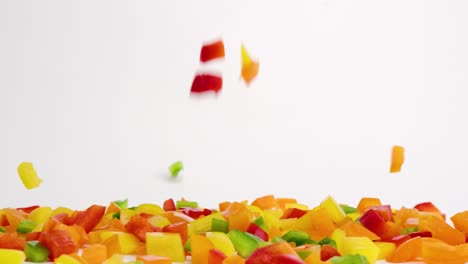 Brightly-colored-red,-green,-yellow-and-orange-diced-bell-pepper-fresh-vegetable-pieces-falling-down-onto-white-table-top-and-bouncing-into-a-pile-in-slow-motion