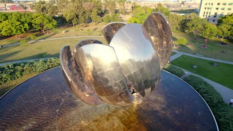 Tilt-down-aerial-view-of-the-stainless-steel-petals-of-the-Floralis-Generica-being-illuminated-by-the-sun's-rays,-round-pool
