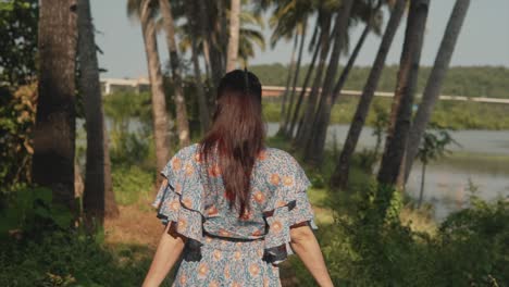 Back-view-of-young-woman-walking-on-a-forest-trail-road-by-a-lake-in-a-floral-print-dress
