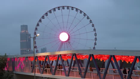 Nighttime-view-of-a-pedestrian-bridge-decorated-with-Chinese-red-lanterns-as-people-walk-through-it-while,-in-the-background,-there-is-a-Ferris-wheel-in-Hong-Kong