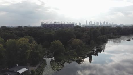 Aerial-drone-shot-of-warsaw-stadium,-with-parallax-effect-with-city-skyline,-lake-and-forest-in-the-background