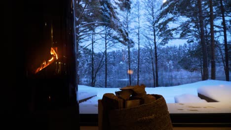 Beautiful-view-of-snow-covered-trees-in-the-forest-and-the-lake-through-the-window-of-a-house-with-a-burning-fireplace-in-cold-winter-evening