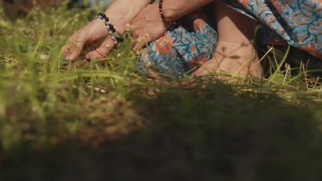 A-close-up-pan-shot-of-the-feet-of-a-Caucasian-female-wearing-a-floral-summer-dress,-crouched-on-her-haunches-picking-at-blades-of-grass-in-a-garden-on-a-beautiful-day-outdoors