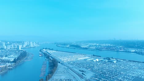 car-dealership-vehicle-storage-4k-drone-flyover-island-filled-in-rows-overlooking-New-Westminster-port-of-trade-dock-for-Import-export-lot-on-a-sunny-day-in-Vancouver-BC-Canada-logs-floating-on-river
