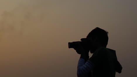 Silhouette-Eines-Jungen-Mannes,-Der-Mit-Einer-DSLR-Kamera-Gegen-Den-Orangefarbenen-Himmel-Fotografiert