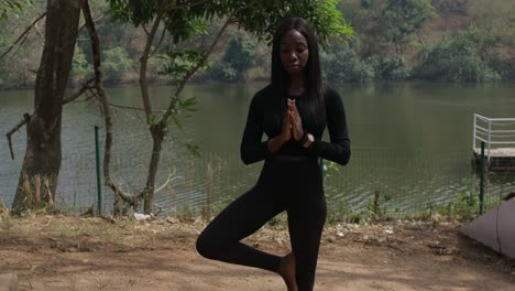 A-lady-during-yoga-in-the-tree-pose-by-a-lake,-static-wide-shot