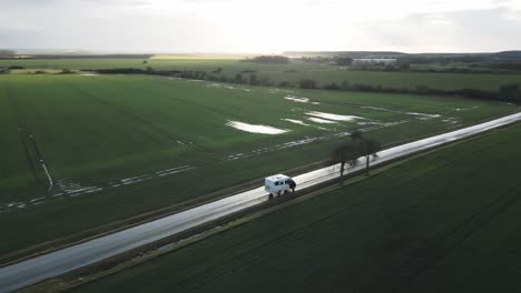 Aerial-drone-view-of-campervan-driving-road-between-green-fields,-flat-landscape,-sunny-day-after-rain
