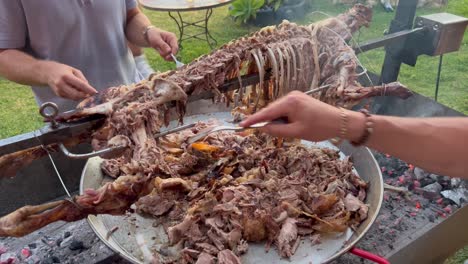 Men-hands-cutting-a-whole-lamb-with-forks-like-savages-on-a-spit-metal-stick-with-golden-crispy-skin,-lamb-bbq-grill-summer-garden-party,-guys-cooking,-4K-shot