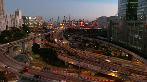 Aufsteigende-Luftaufnahme-Des-Verkehrsnetzes-Im-Bezirk-Kwai-Chung-Nach-Sonnenuntergang---Industriehafen-Von-Hong-Kong-City-Im-Hintergrund