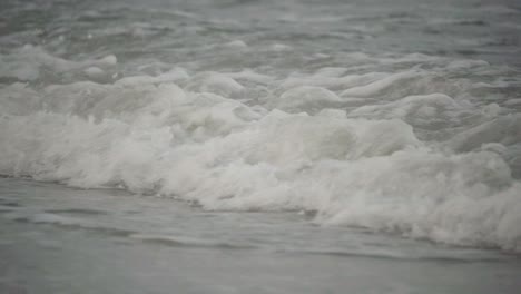 Closeup,-ocean-tide-crashing-onto-beach-shore-on-a-gloomy-day-in-slow-motion