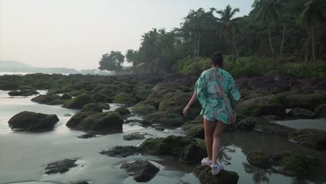 Beautiful-young-woman-walking-on-mossy-rocks-on-a-tropical-beach-with-palm-trees-in-the-background