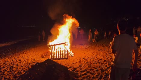 Traditional-bonfire-festival-at-the-beach-at-the-San-Juan-celebration-in-Marbella-Spain,-people-drinking,-friends-and-family-enjoying-fun-party-during-summer,-big-burning-fire-and-hot-flames,-4K-shot