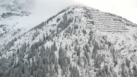 Aerial-zoom-shot-of-snow-packed-mountain-top-with-snow-retention-and-trees