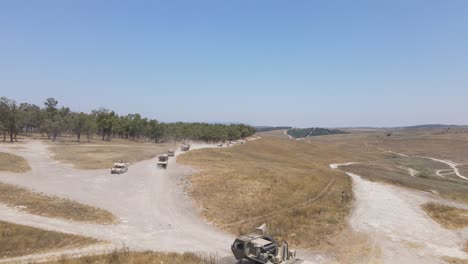 Israel-Army-infantry-squad-soldiers-on-vehicles-driving-through-green-field-at-training-ground-country-road,-Aerial-Tracking-shot