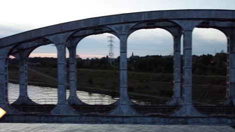 Old-steel-railway-bridge-over-river-surrounded-by-meadows,-aerial-side-fly