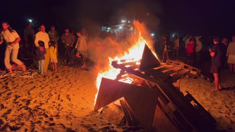 Traditional-bonfire-festival-at-the-beach-at-the-San-Juan-celebration-in-Marbella-Spain,-friends-and-family-enjoying-a-fun-party-during-summer,-big-burning-fire-and-hot-flames-at-night,-4K-shot