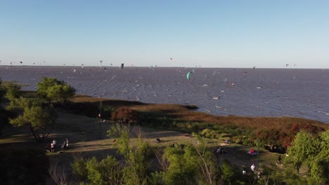 Luftbahnaufnahme-Vieler-Kitesurfer-Am-Fluss-Mit-Bäumen-An-Der-Küste-In-Vicente-Lopez-In-Buenos-Aires