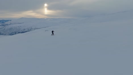 Lone-skier-in-snowy-winter-mountain-walking-purposefully-towards-the-top---Afternoon-aerial-with-cloudy-sky-and-deep-valley-background---Norway