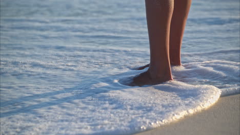 Slow-motion-of-a-pair-of-tanned-thin-woman-legs-standing-on-the-sand-having-the-waves-splashing-on-her-at-the-beach
