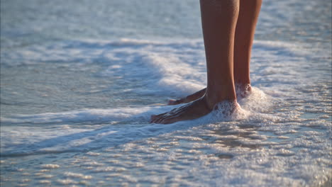 Slow-motion-of-a-pair-of-tanned-woman-legs-standing-on-the-beach-with-the-water-splashing-on-her