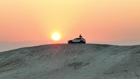-Man-sits-on-a-desert-hill-overlooking-a-sunrise-in-on-top-of-a-car