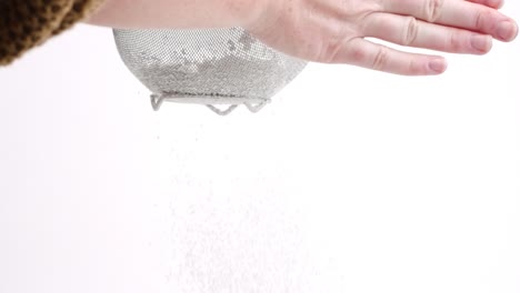 White-female-hand-sifting-powdered-sugar-in-sieve-on-white-backdrop-in-slow-motion