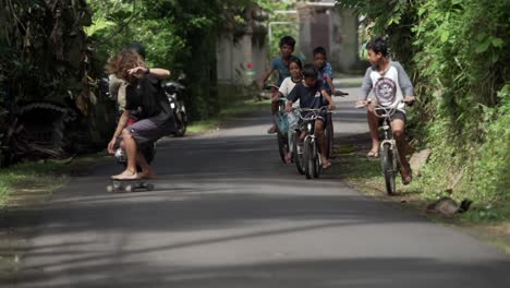 Indonesian-kids-follow-European-tourist-skating-barefoot-down-the-street,-Slow-motion