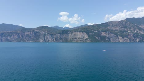 A-panoramic-aerial-shot-of-the-majestic-mountains-looming-over-Lake-Garda-in-the-charming-town-of-Malcesine,-Verona,-Italy-on-a-picturesque-day-with-a-clear-blue-sky-and-fluffy-clouds