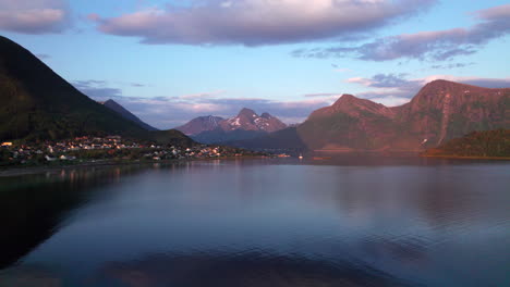 Aerial-panning-shot-of-a-small-town-nestled-in-a-fjord-along-the-cost-of-Norway-during-a-beautiful-sunset-with-mountains-and-clouds