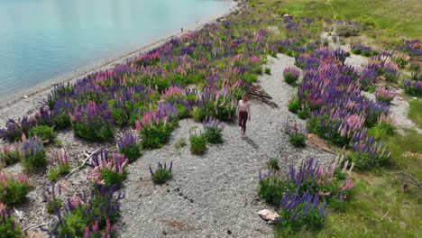 Fit-woman-in-sports-clothing-walking-on-pebble-lake-shore-with-Lupin-flowers