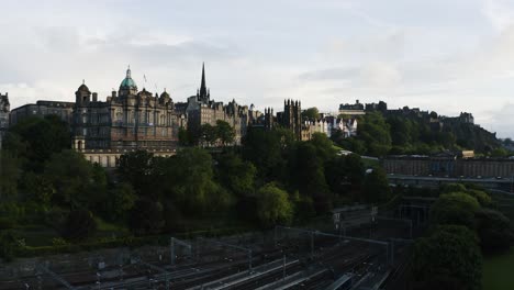 Rising-aerial-shot-featuring-Waverley-Train-Station,-Museum-on-the-Mound,-and-The-Hub-in-Edinburgh,-Scotland