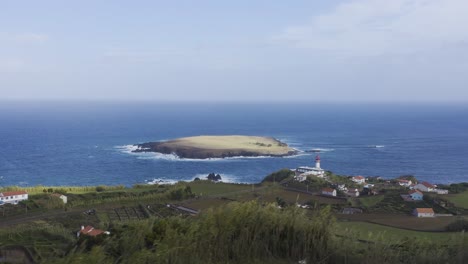 Drone-view-of-a-rural-coastal-village-with-a-lighthouse-and-an-island-in-the-Atlantic-ocean,-cloudy-sky-in-Topo,-São-Jorge-island,-the-Azores,-Portugal