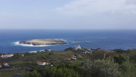 Drone-view-of-a-rural-coastal-village-with-a-lighthouse-and-an-island-in-the-Atlantic-ocean,-cloudy-sky-in-Topo,-São-Jorge-island,-the-Azores,-Portugal