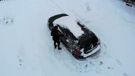 Man-sweeps-snow-off-the-black-car-after-heavy-snowfall,-aerial-view