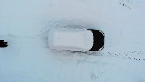 Driver-walks-towards-the-snow-covered-black-car-after-blizzard-in-winter,-aerial-view