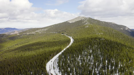 Aerial-reveal-backward-of-sunny-and-snow-covered-back-roads-in-the-Colorado-mountains-surrounded-by-bright-green-pine-tree-forests-with-blue-skies