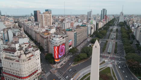 Aerial-pan-right-of-Obelisk-in-the-middle-of-9-de-Julio-avenue-traffic-and-metrobus-station,-Buenos-Aires
