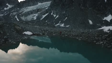Male-Hiker-Standing-in-Awe-of-the-Rugged-Alaskan-Mountain-Range-with-Epic-Reflection-Shot-on-Drone-from-a-distance
