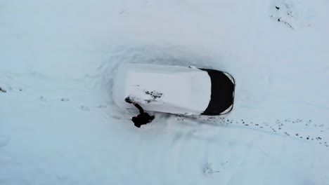 Man-sweeps-out-frozen-snow-layer-from-black-car-after-winter-snowfall,-aerial