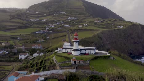 Drone-view-of-a-lighthouse-in-an-rural-coastal-village,-cloudy-sky-in-Topo,-São-Jorge-island,-the-Azores,-Portugal