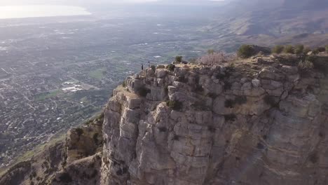 Aerial-orbit-shot-of-two-hikers-standing-on-a-mountain-peak-in-Provo,-Utah-with-drone-panning-to-reveal-the-town-below