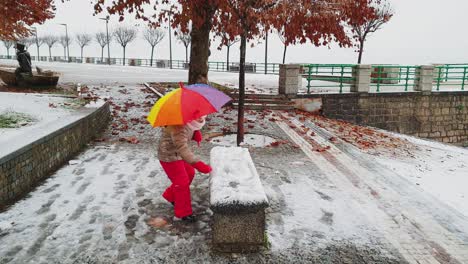 Little-child-girl-with-umbrella-of-many-colors-plays-kicking-snowball